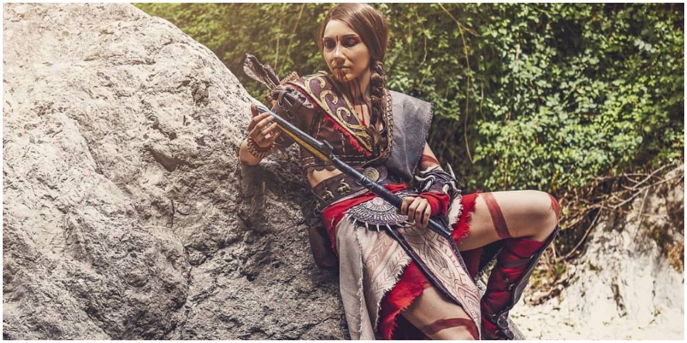 A cosplay of Kassandra resting on rocks and looking at her weapon
