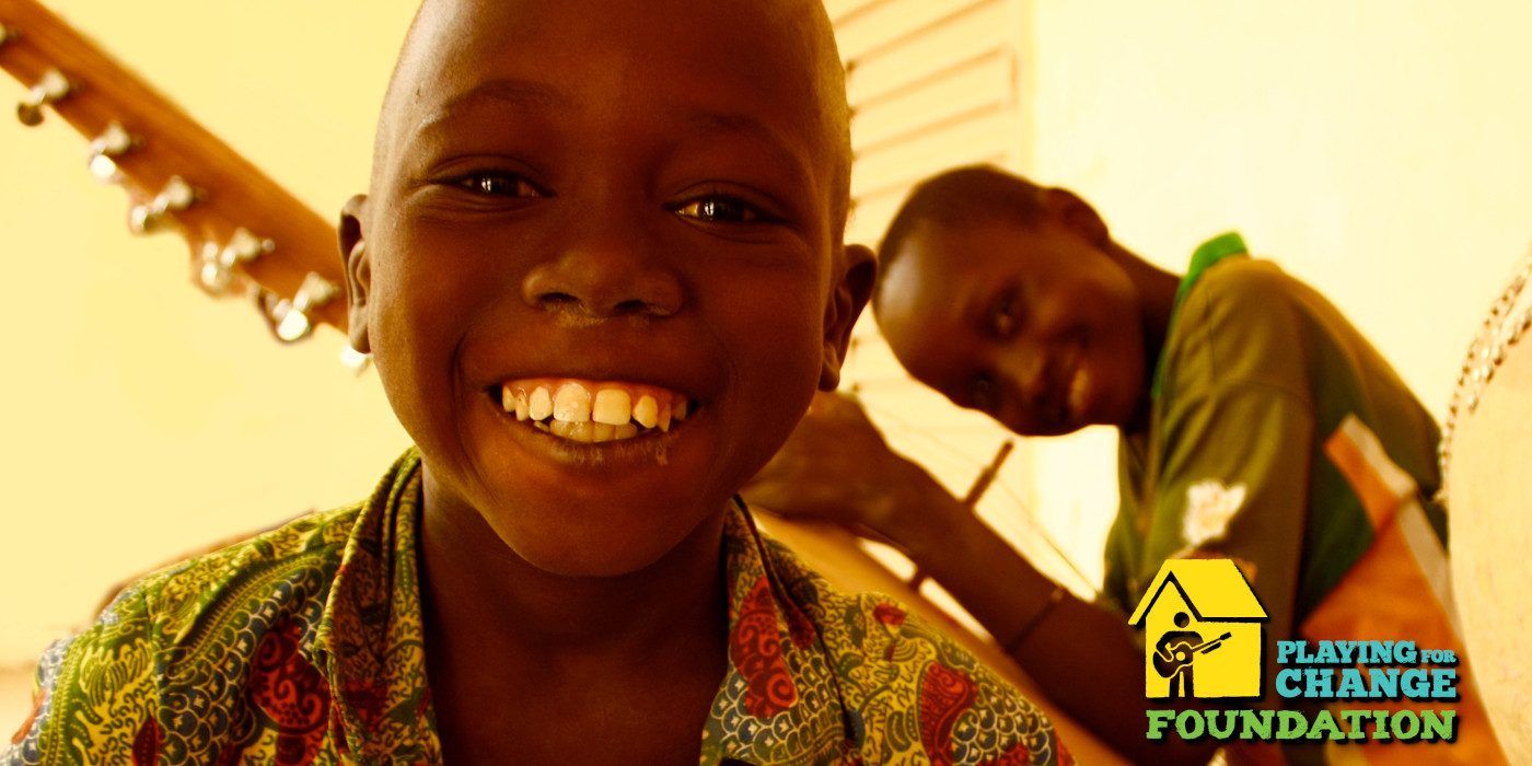 Two boys grin while holding instruments. Logo in lower right for the Playing For Change Foundation.