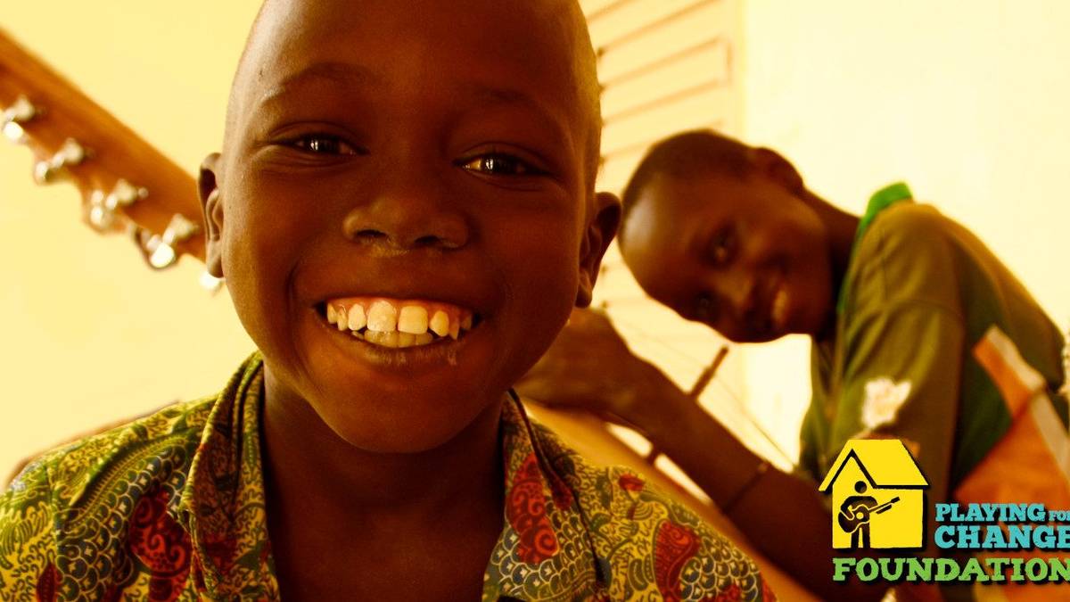 Two boys grin while holding instruments. Logo in lower right for the Playing For Change Foundation.