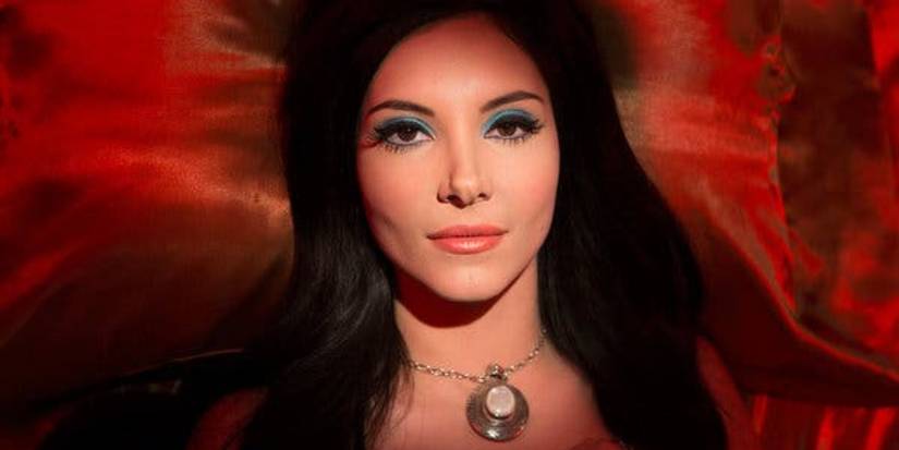 The Love Witch still of a woman laying on red satin pillow wearing blue eyeshadow and silver necklace