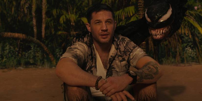 tom hardy in a Hawaiian shirt sitting on a beach with the venom symbiote head next to him