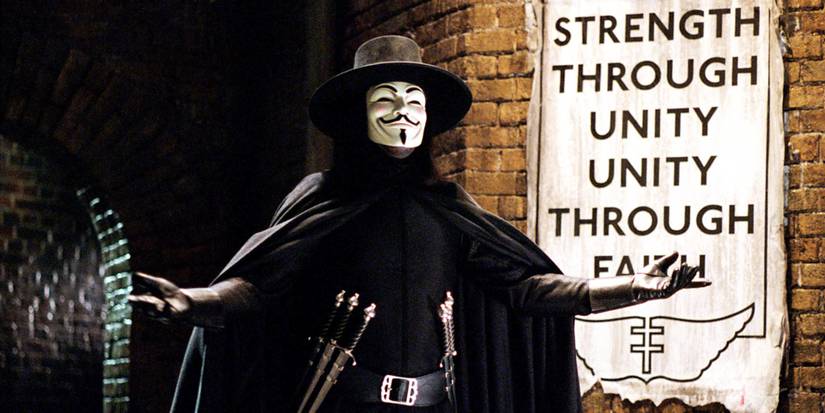 V, played by Hugo Weaving, dressed in a Guy Fawkes mask and carrying knives, stands in an alley in V For Vendetta
