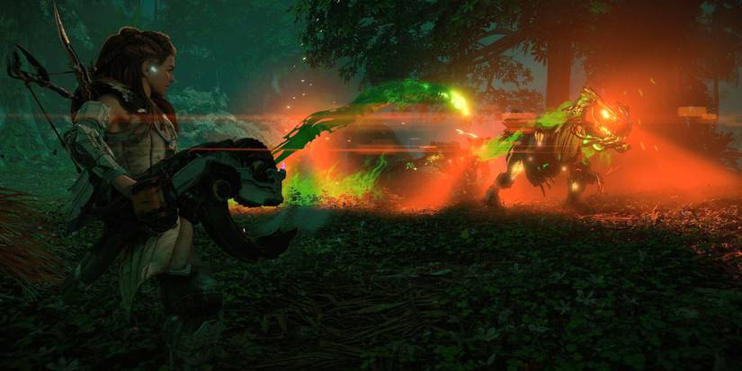 Aloy holds a large weapon and fires a glowing, green projectile that trails green slime at a mechanical raptor with glowing red eyes in some nearby trees