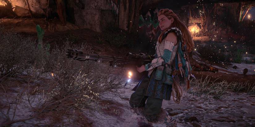 aloy walking through a desert camp with a large, long-barreled gun in her hands