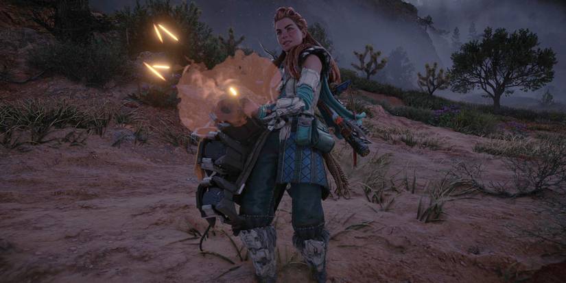 Aloy, in a green and white outfit with metal plates over her shins and forearms, holds a heavy piece of machinery that has a cloud of smoke and floating embers around the nozzle 