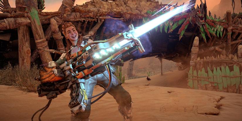 aloy, with a grin on her face and her tongue sticking out, holds a large gun with a blue, glowing beam shooting out of the nozzle