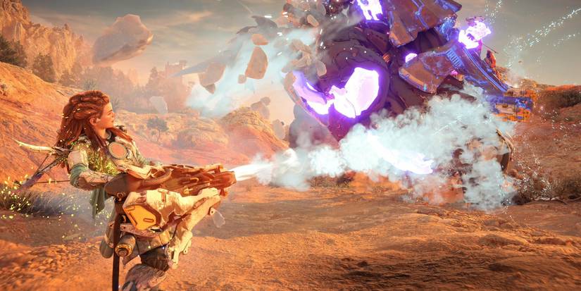 aloy holding a large gun that's shooting out a steady stream of light blue mist at a large, turtle-like machine a short distance away 