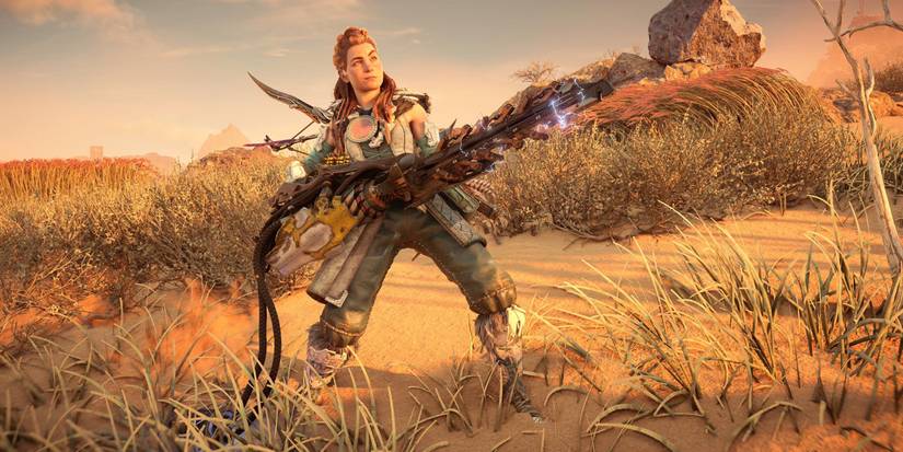 aloy holding a large gun with a long, sparking barrel while standing in a desert with short grass and reeds nearby 