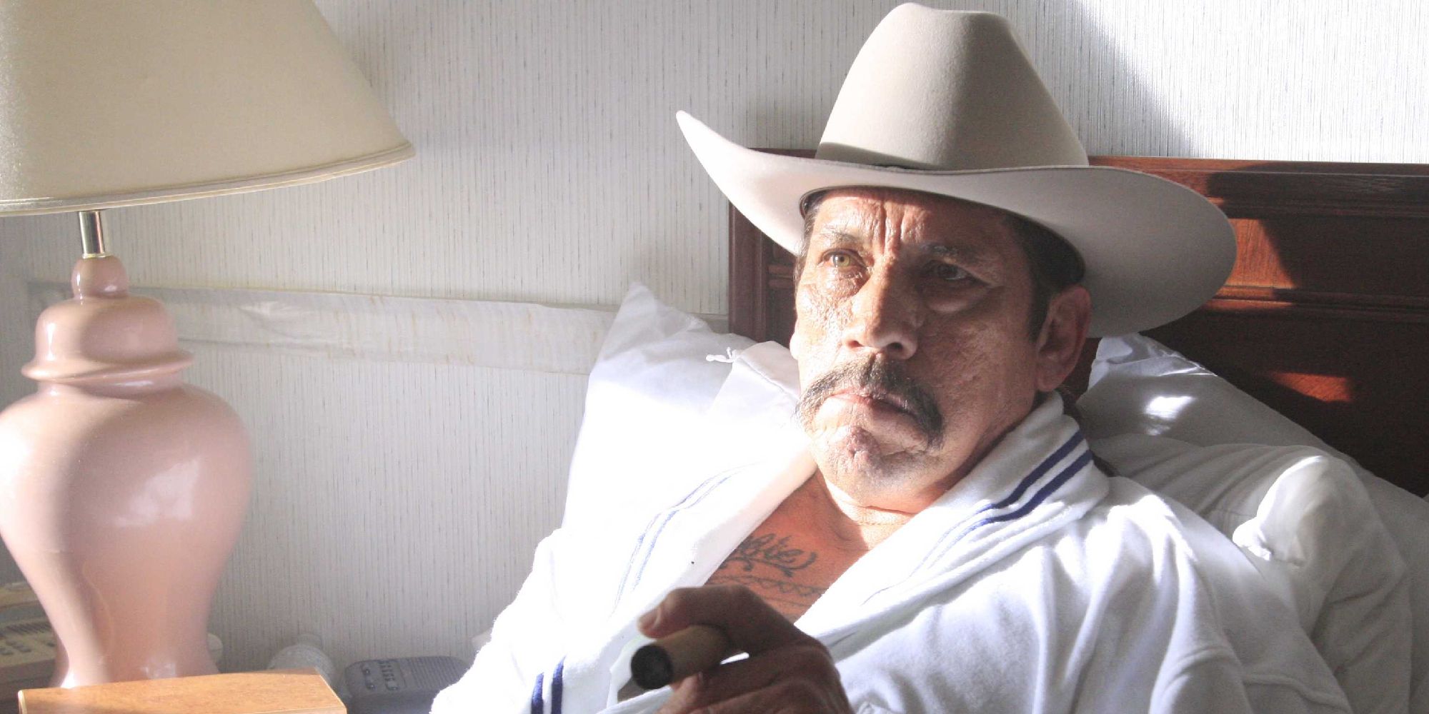 Danny Trejo as Tortuga sitting on a hotel bed in a cowboy hat and robe, smoking a cigar