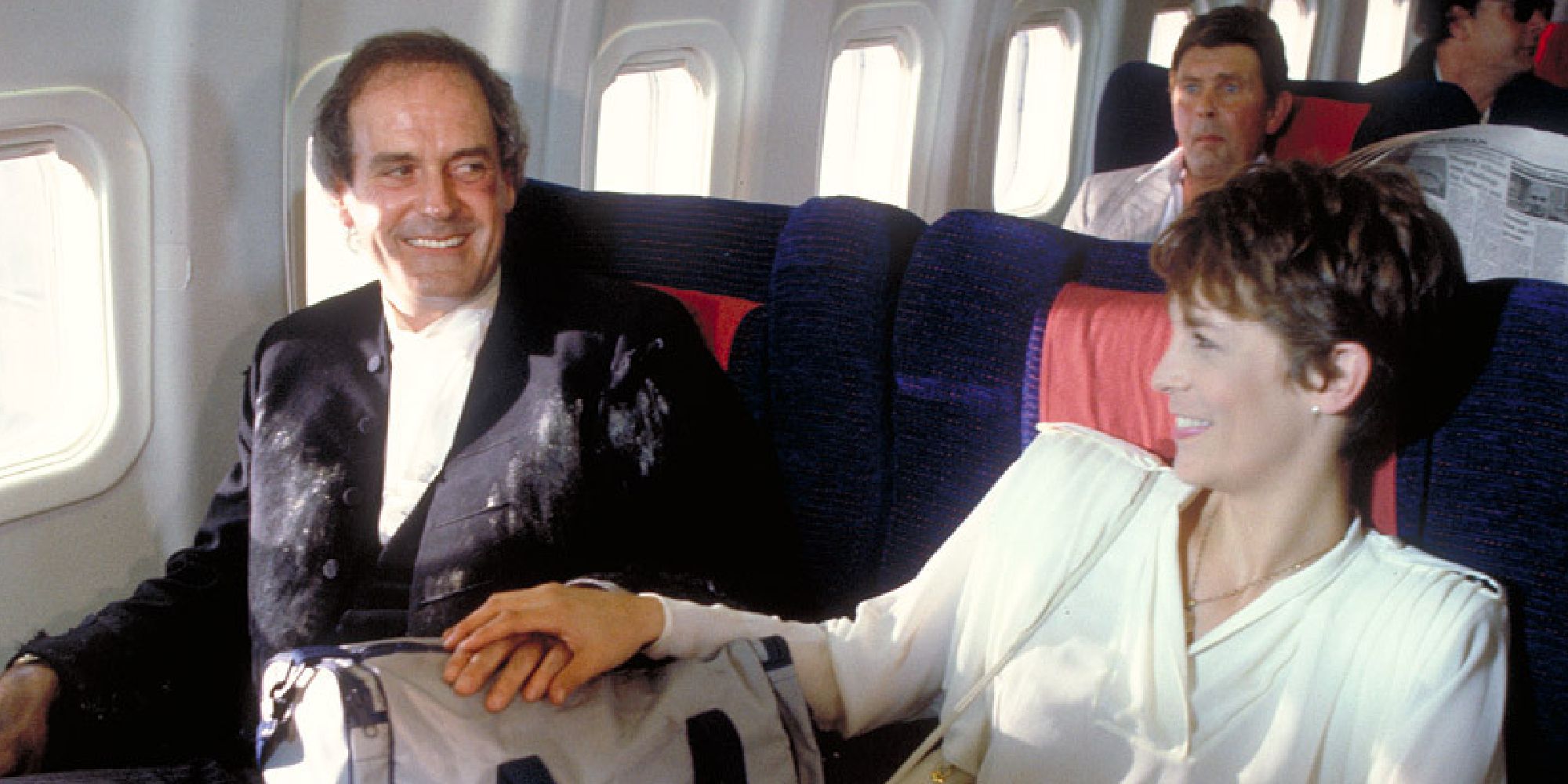 John Cleese and Jamie Lee Curtis holding hands on a plane with a duffel bag in A Fish Called Wanda