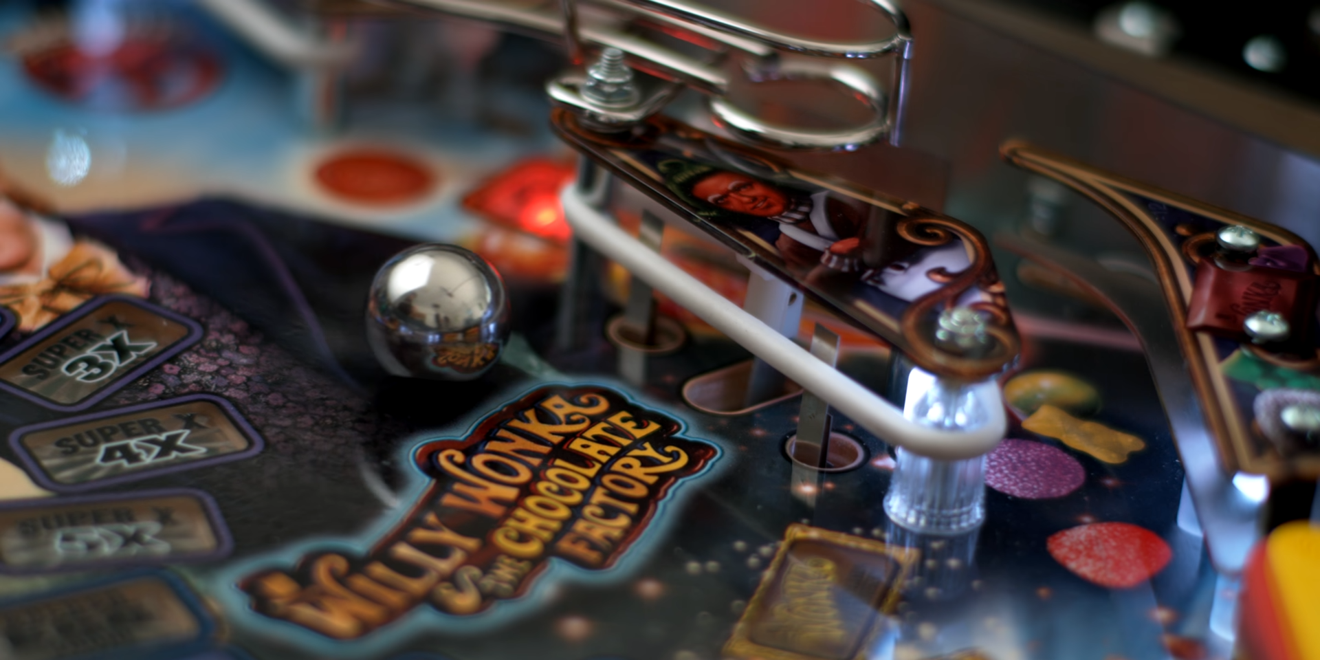 A ball bearing rolling towards a bumper on a brightly-colored pinball machine. The words "Willy Wonka and the Chocolate Factory" appear in a swirly font on the table. Image credit: The Slow Mo Guys YouTube