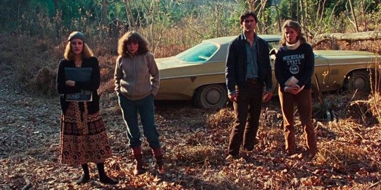 Four of the 1981 Evil Dead's cast members stand on leafy ground in front of an Oldsmobile car.