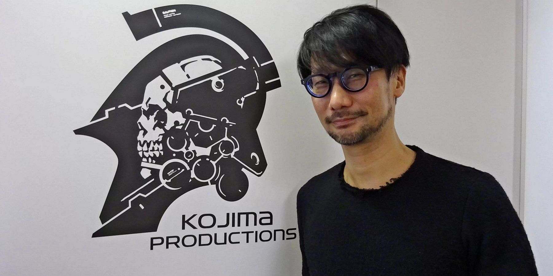 Photo of Hideo Kojima in front of the Kojima Productions logo.