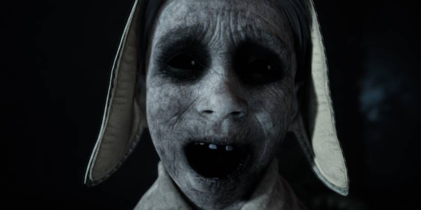 Close up of a grey, cracked visage of a ghost girl, her mouth wide open and eyes sorrowful as though she wails tagically. 