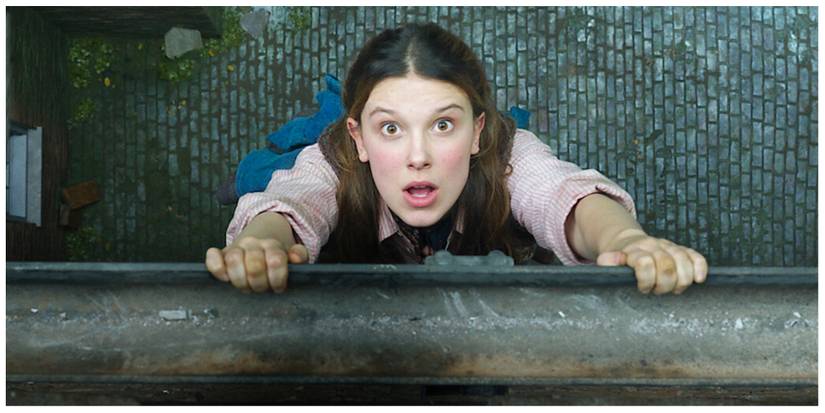 Enola Holmes hanging from the gutter from a house looking shocked