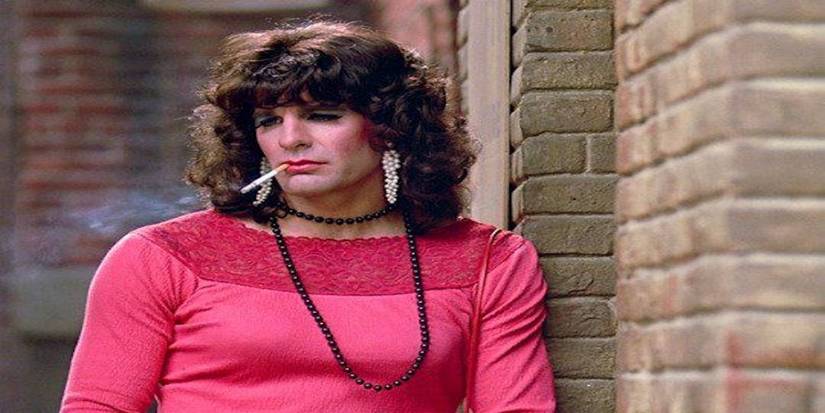 Sam is dressed as a woman in a pink top and bead necklace smoking a cigarette as an undercover Police Detective Jake Rawlins in M.I.A