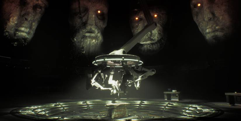 Four large statue faces overlook a device with many arms in the center of a dark, futuristic room.