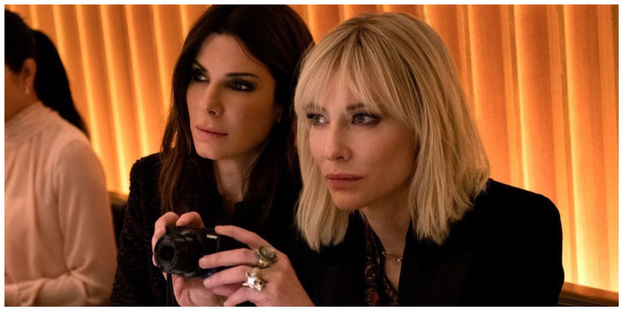 Cate Blanchett in Ocean's 8 taking pictures with a camera, whilst stood next to Sandra Bullock. They are both looking at something off screen.