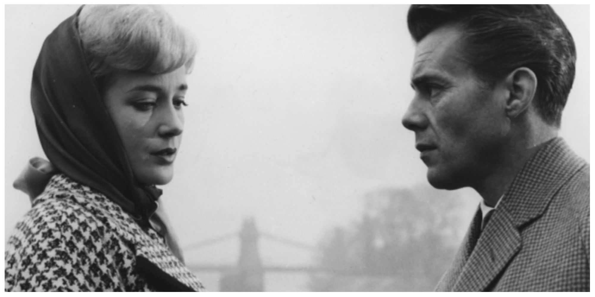 Laura (Sylvia Syms, left) and Melville Farr (Dirk Bogarde) from the film looking at each other in conversation outside next to the River Thames. Hammersmith Bridge can be seen in the background of the shot.