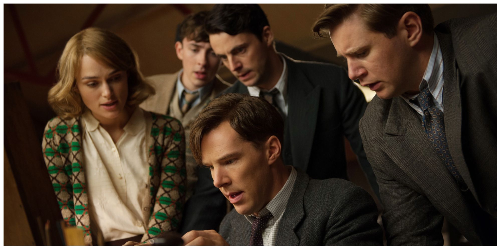 Benedict Cumberbatch as Alan Turing sat looking at something on the table surrounded by supporting cast. They all look shocked.