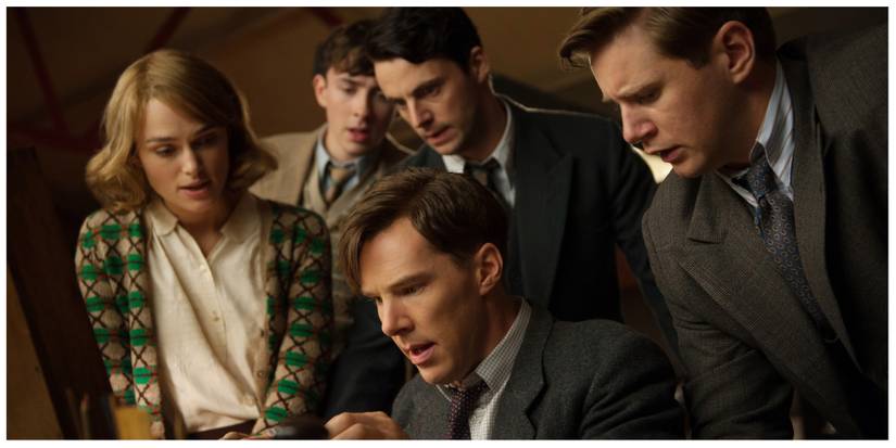 Benedict Cumberbatch as Alan Turing sat looking at something on the table surrounded by supporting cast. They all look shocked.