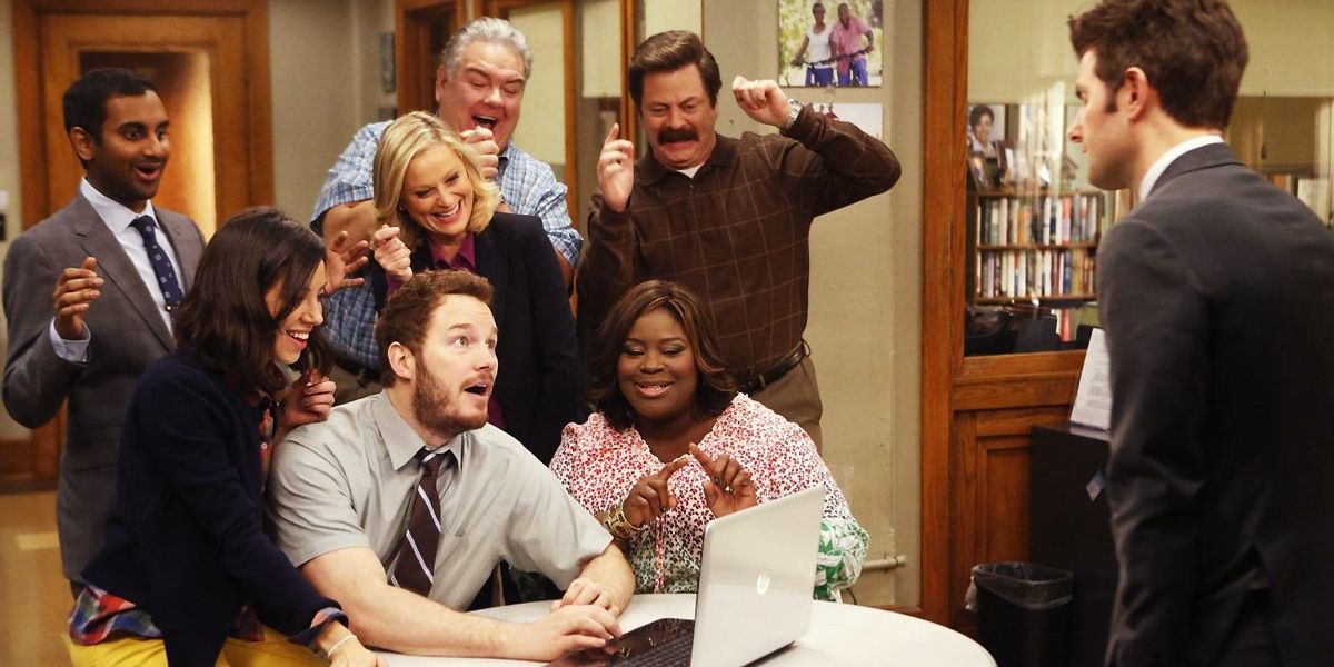 The cast of Parks and Recreation gather around a laptop and cheer