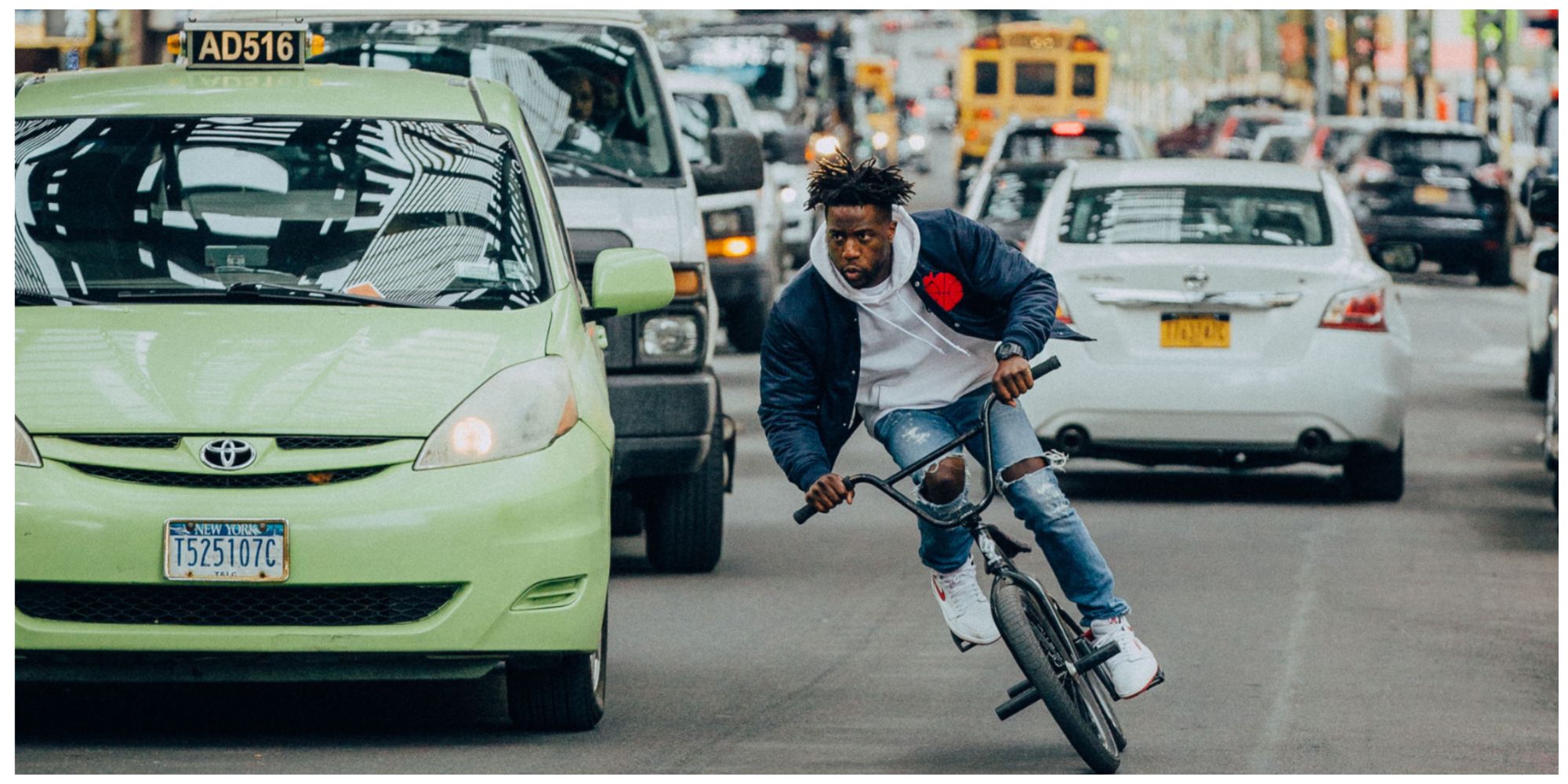 Nigel Sylvester riding his BMX through traffic in ripped jeans, a white hoodie and black jacket