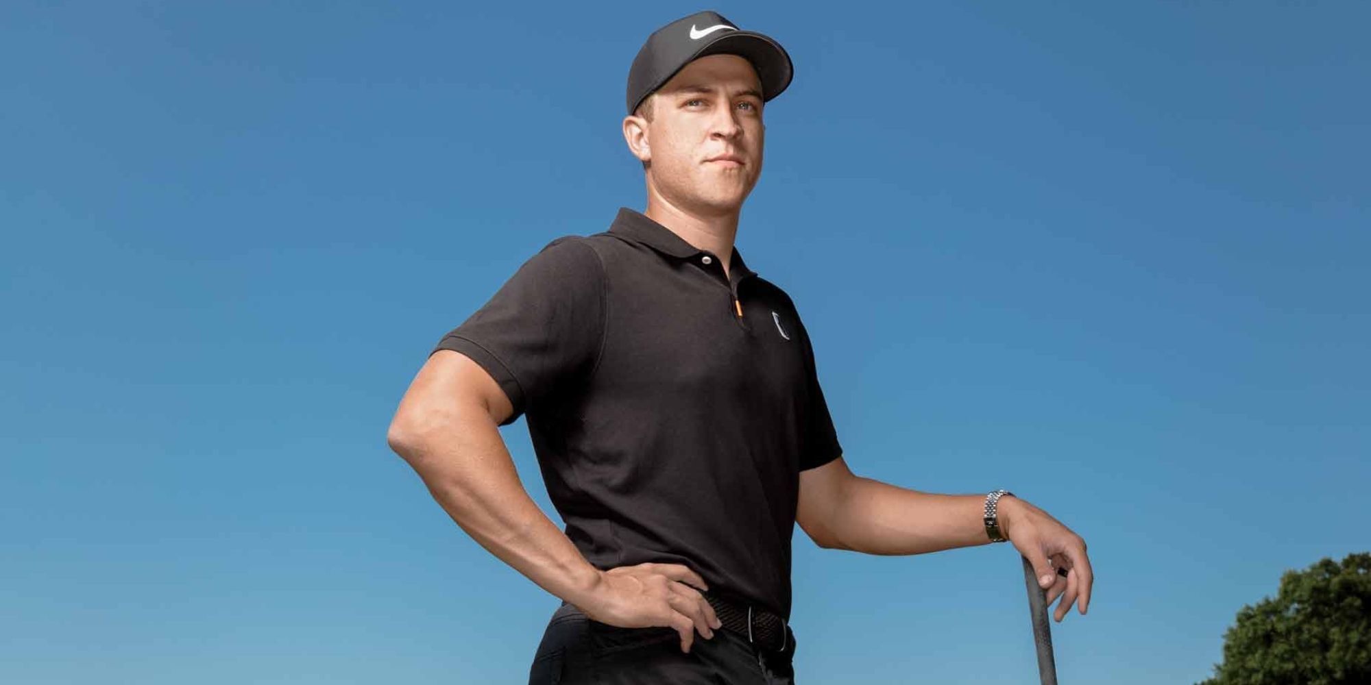 PGA Golfer Cameron Champ posing in a black polo shirt and black Nike cap. He is leaning on his golf club.