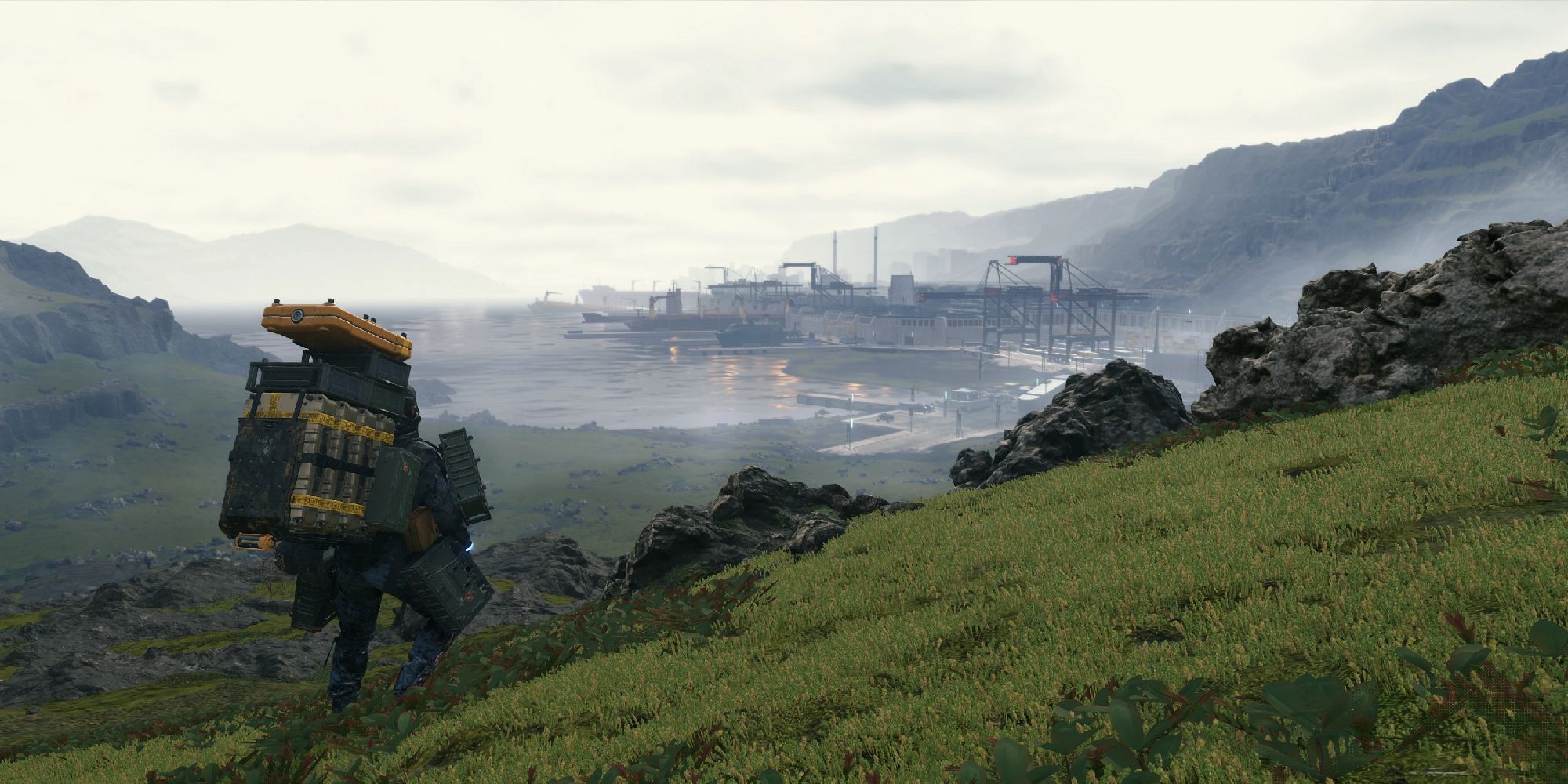 The protagonist stands on a grassy hill overlooking a structure in the distance in Death Stranding