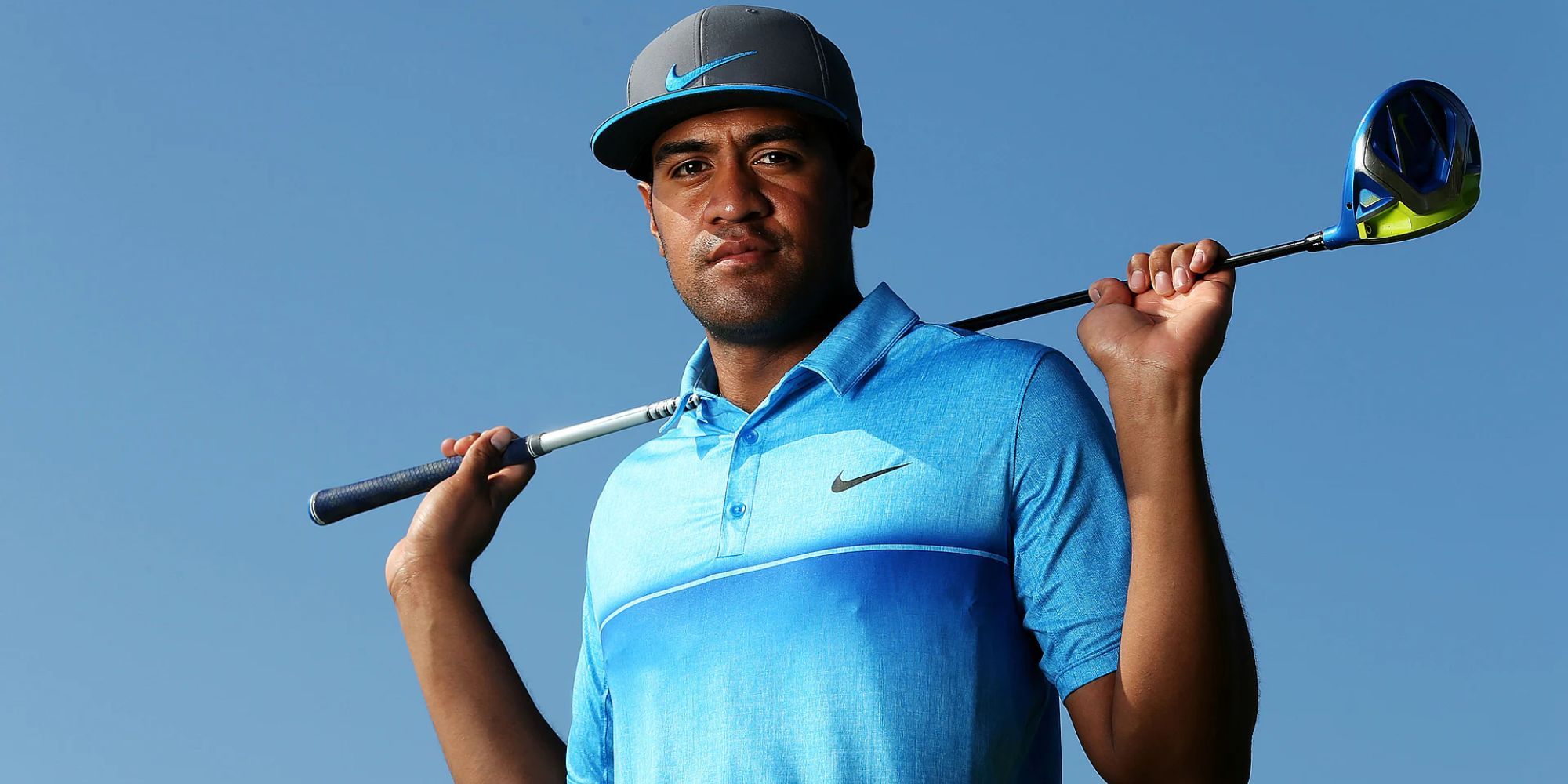 PGA Golfer Tony Finau posing for a photo in a light blue polo shirt and black cap. He is holding his golf club across his shoulders.