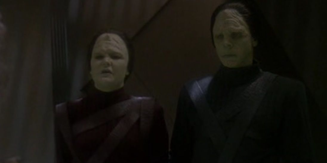 Two pale green aliens, a woman and a man, wearing dark clothes in a dim cell.