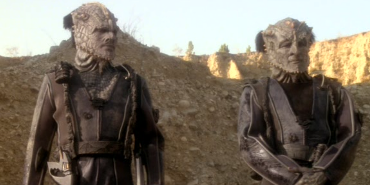 Two alien warriors with blue-gray scales standing against a rocky landscape.