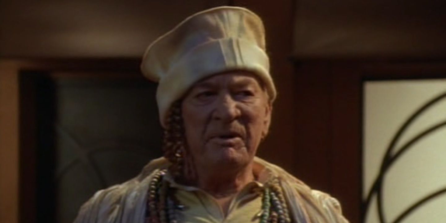 An elderly man in a round cream hat. He is wearing many beads. 