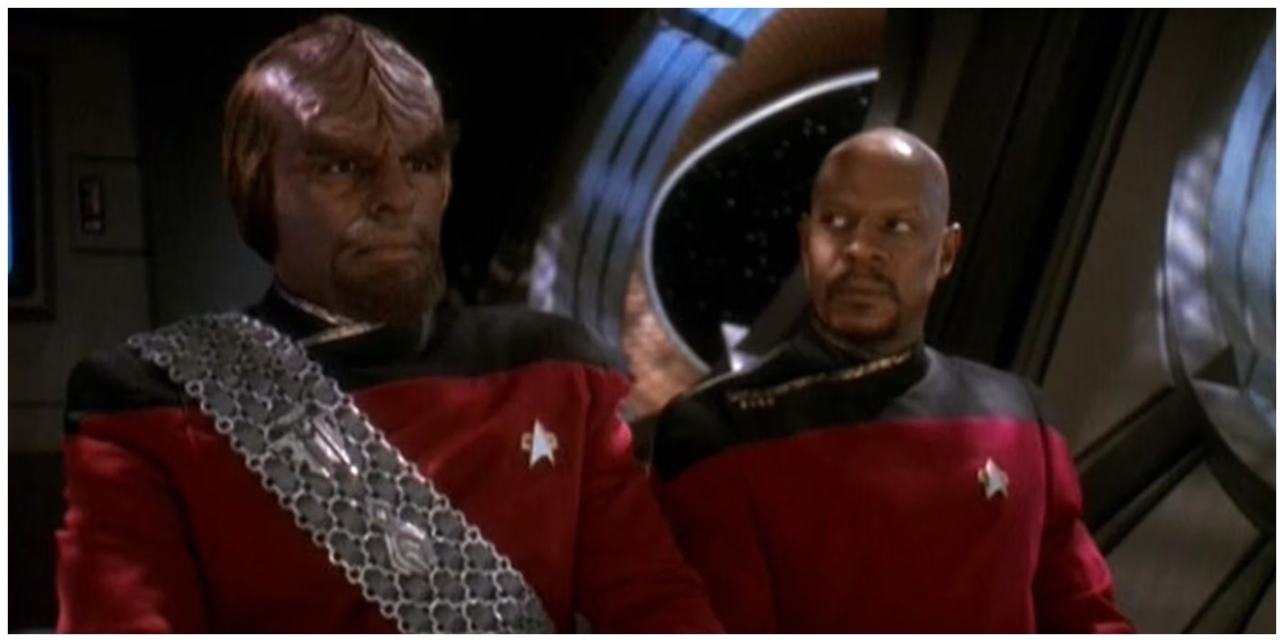 Michael Dorn, a Black man, in Klingon makeup that gives him a ridged forehead from browline to the top of his head and a brown goatee, stands in his red Starfleet uniform and silver warrior's sash next to Avery Brooks, a Black man who is bald with a brown goatee., also wearing a red Starfleet uniform. Avery is looking at Dorn with a serious expression.