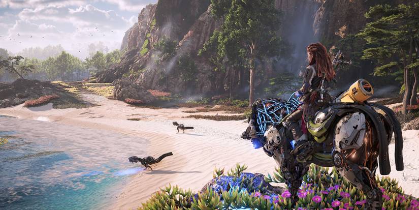 Aloy sits on a robotic mount looking out at a beach with two machine dinosaur-like creatures roaming on it.