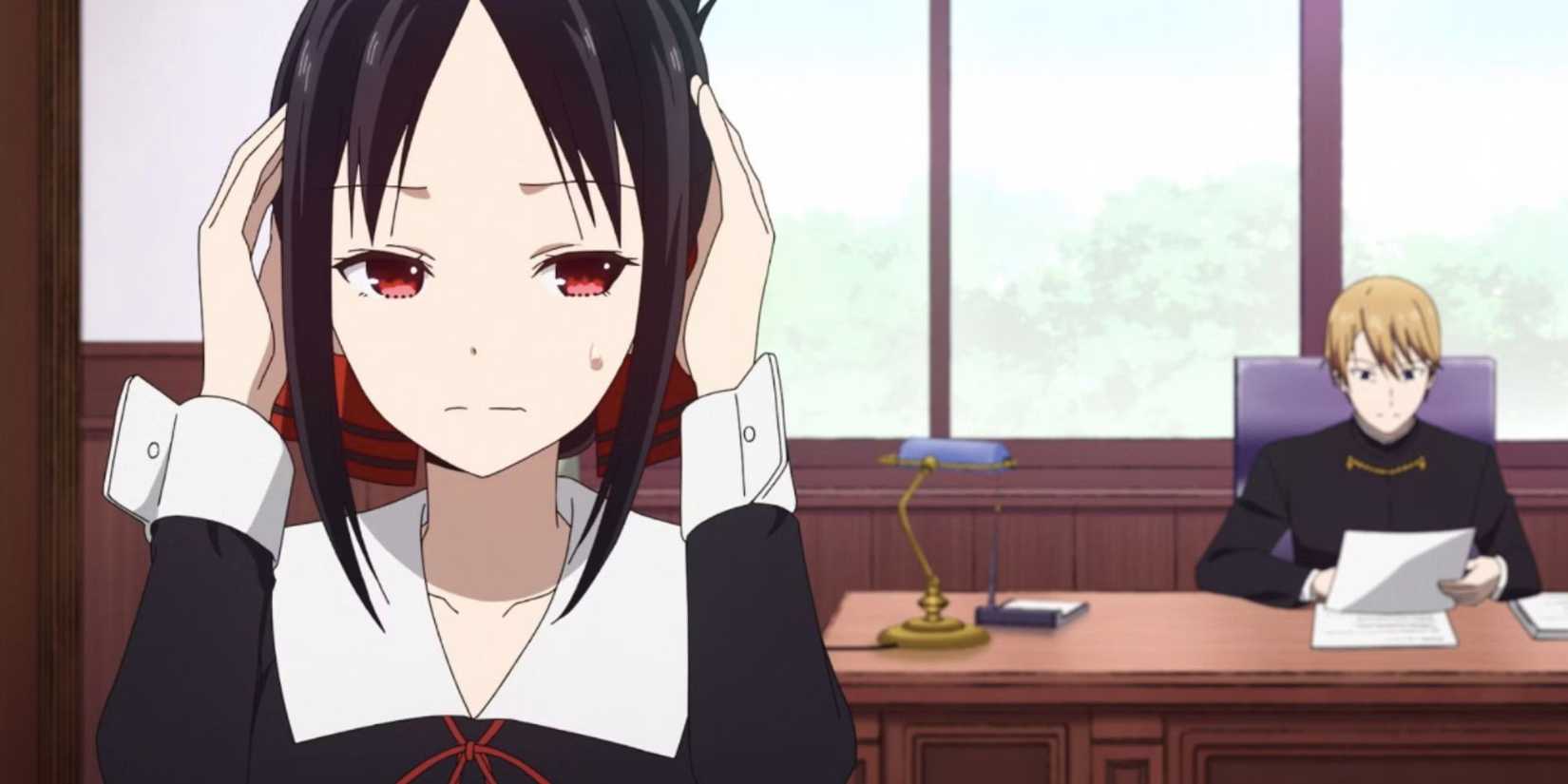 a black haired girl stands in the foreground, a blonde boy sits at a desk in the background