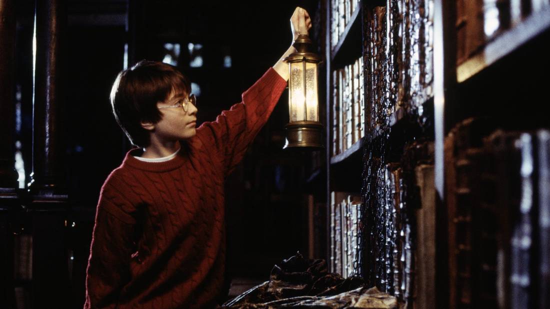 A boy in a red sweater is lifting a lantern to view old books. A cloak is set down infront of him.