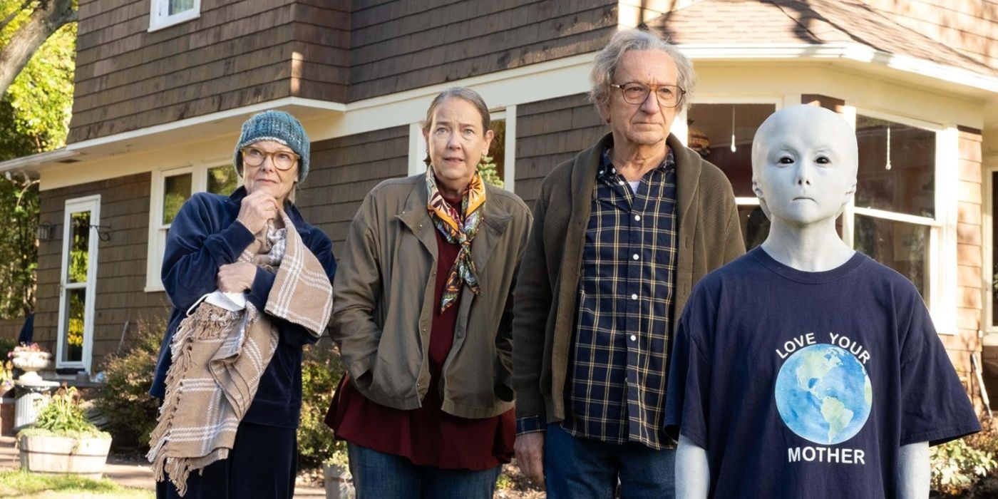 Three elderly people stand in a yard with a small blue alien in front of them