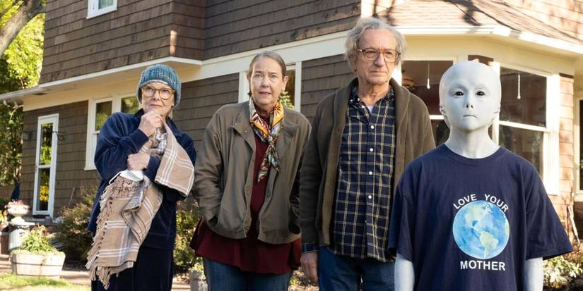Three elderly people stand in a yard with a small blue alien in front of them