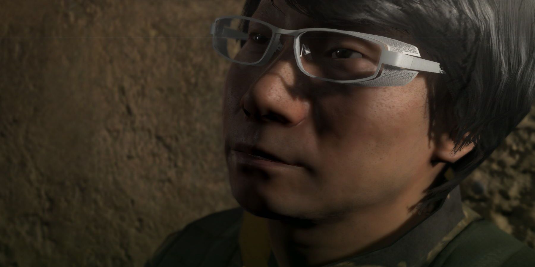 Hideo Kojima sitting in front of a rocky wall in army fatigues