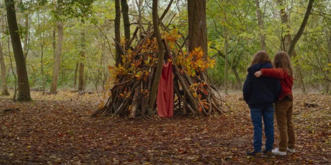 Two little girls embrace and look at a stick fort in the woods