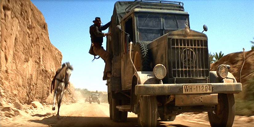 Indiana Jones holds on to the side of a moving truck after jumping onto it from his horse