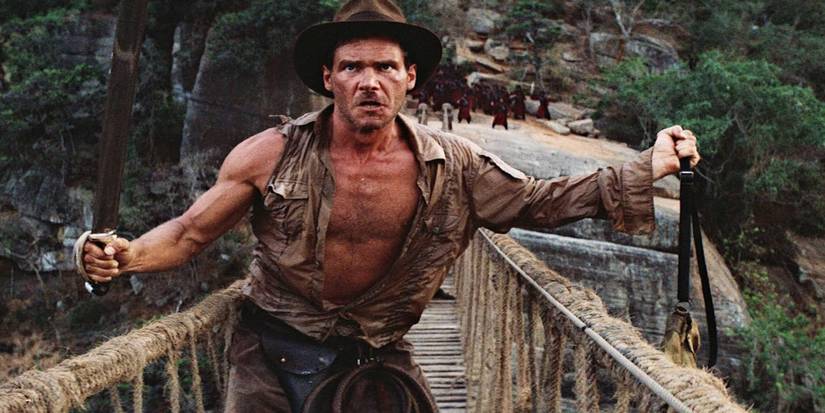 Indiana Jones stands on a wooden bridge dangling sacred stones over the edge, threatening to drop them