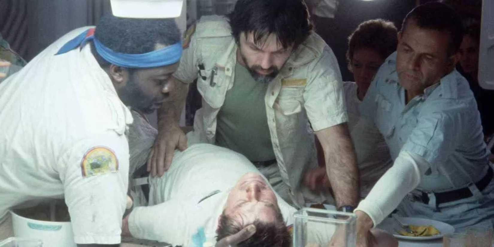 The crew holds down Kane as he cries out in pain