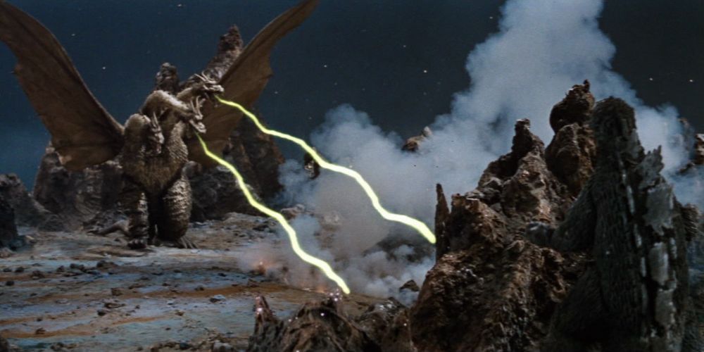 King Ghidorah shoots at Godzilla by he hides behind a rock.