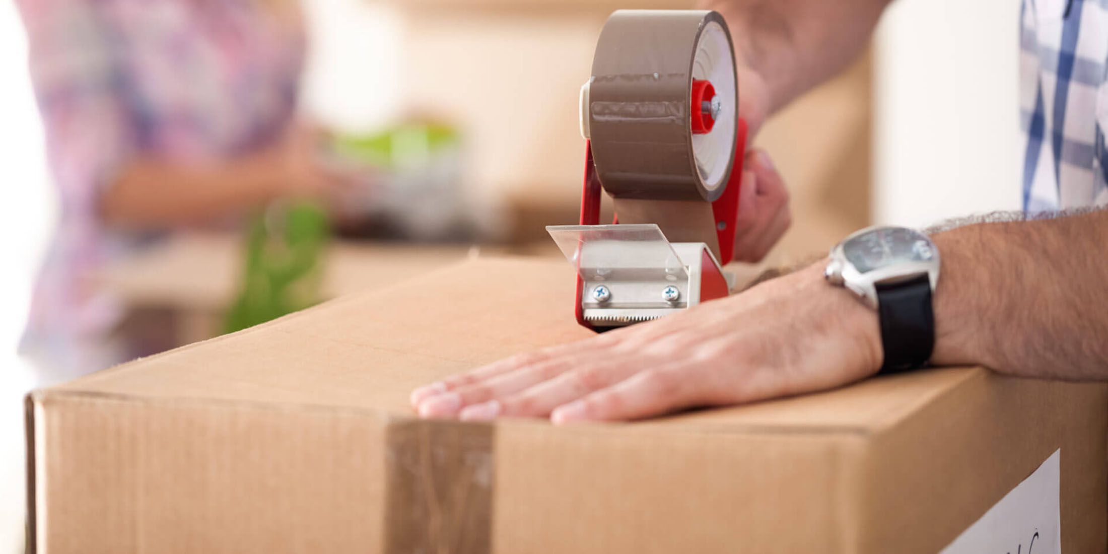 A stock image of someone taping a cardboard box closed while wearing a blue and white flannel shirt.