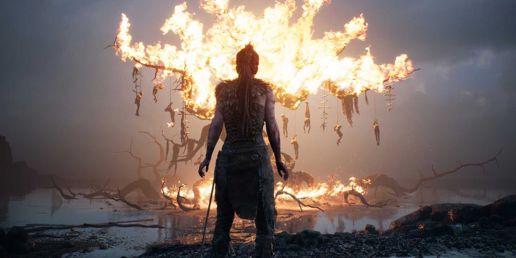Senua stands in front of a  burning tree with hanging bodies in Hellblade Senua's Sacrifice