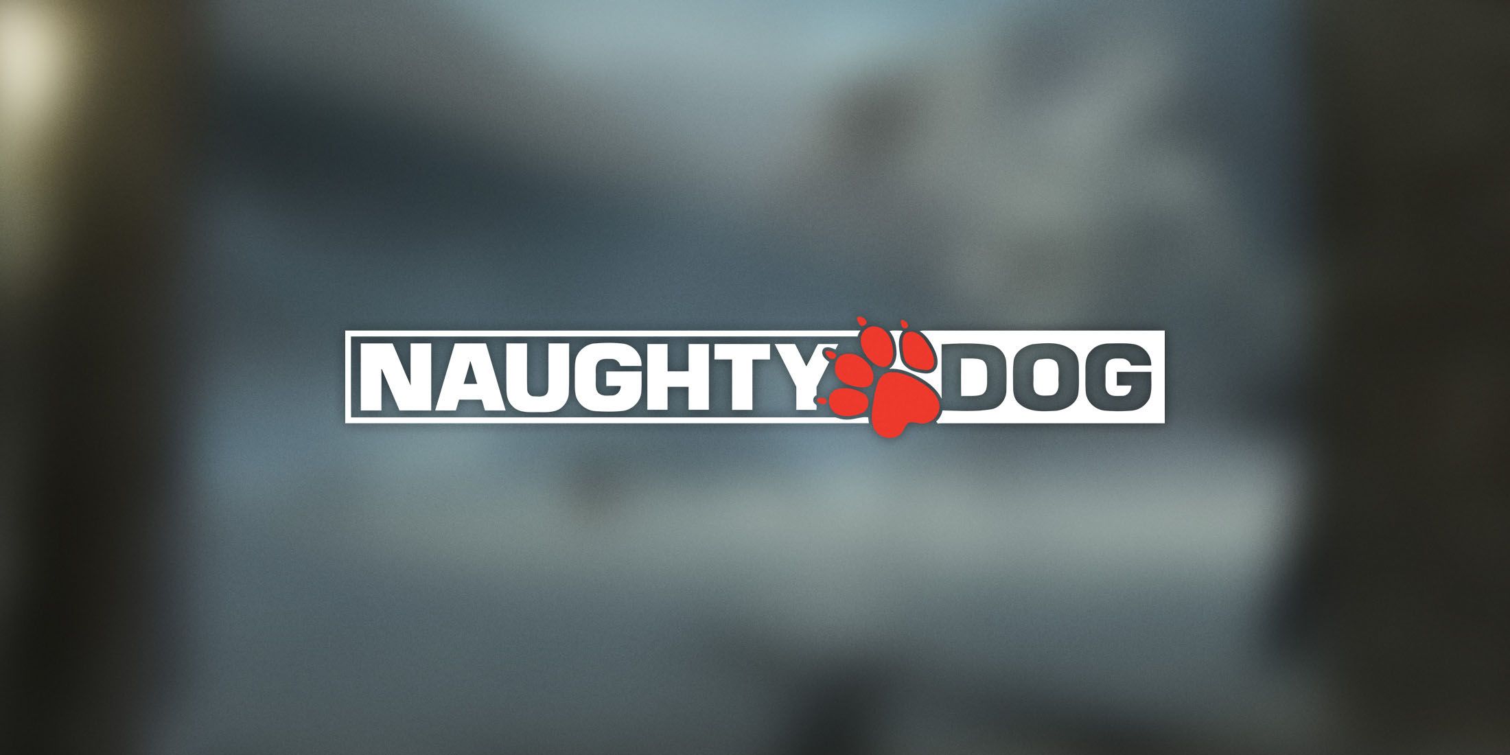 A white and red Naughty Dog logo placed in front of a blurry grey background.