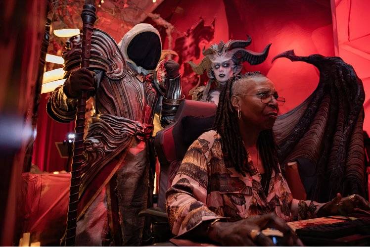 Whoopi Goldeberg sits at a computer desk playing a game under the watch of Diablo 4 characters.