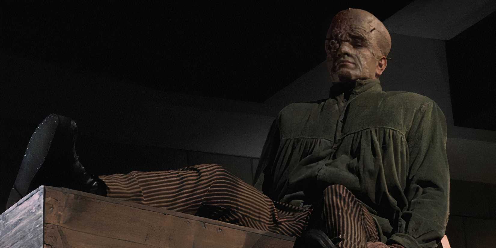 the disfigured creature sits up on the table